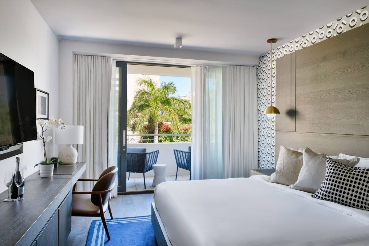 Habitación de hotel con una cama grande, un escritorio con televisión, una silla, un balcón con vistas a palmeras y una pared con diseño.