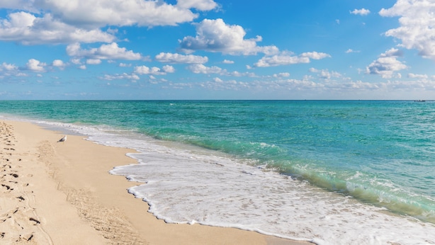 En strand med kristallklart turkost vatten, vit sand och en klarblå himmel med enstaka moln.
