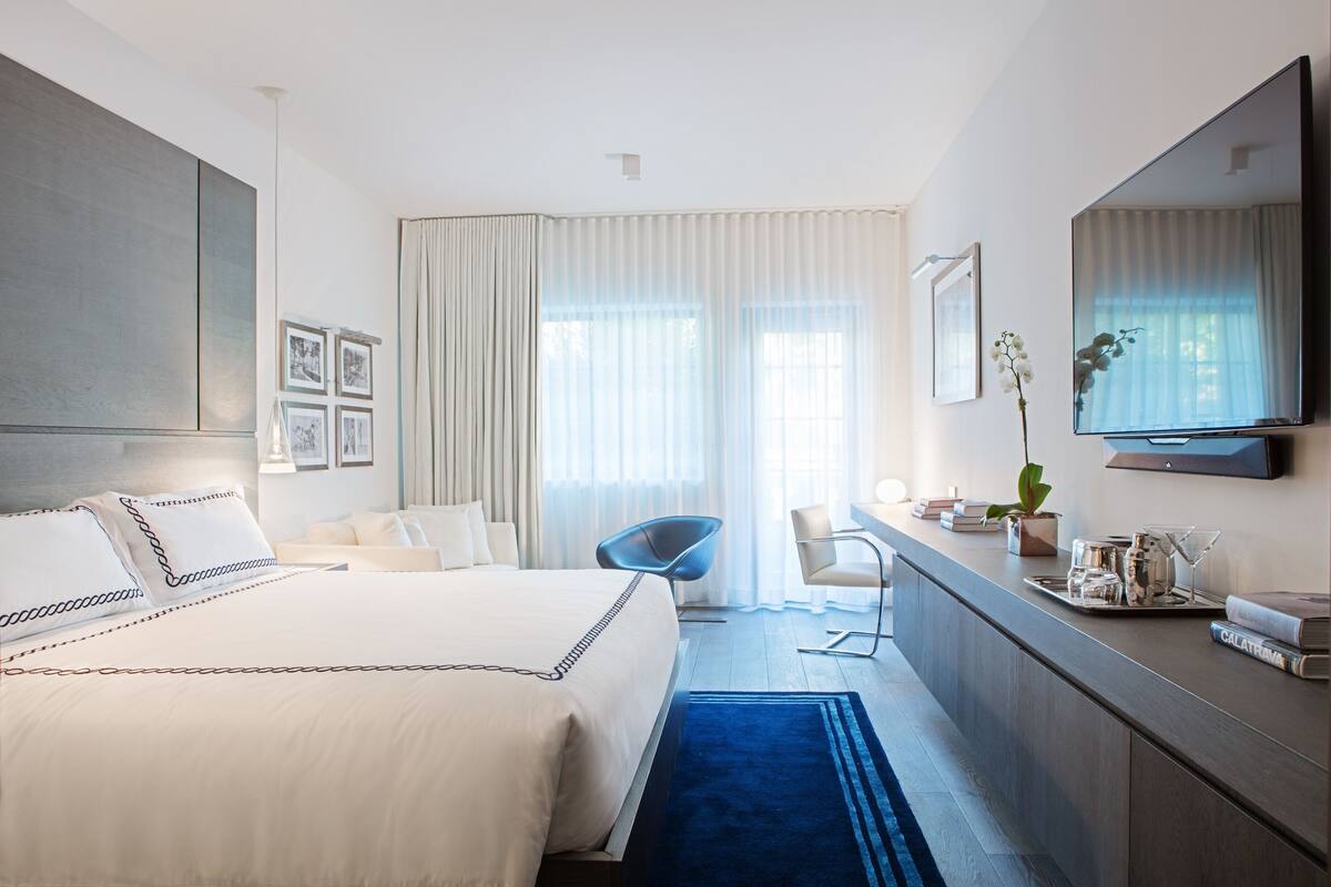 Una habitación de hotel moderna con una cama grande, un escritorio, una silla, un televisor y un amplio ventanal con cortinas.