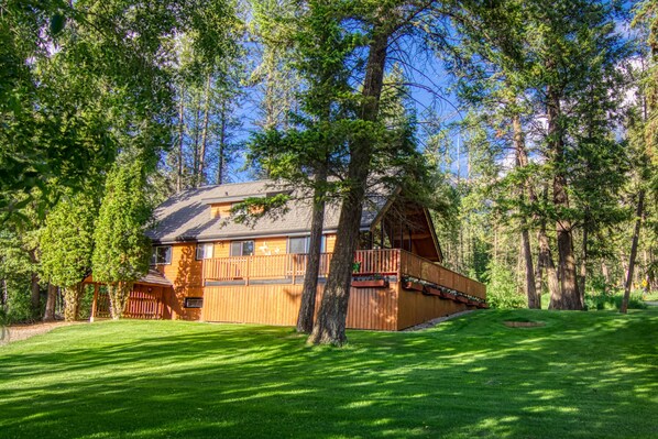 A wooden cabin with a deck, surrounded by trees and a well-maintained lawn.