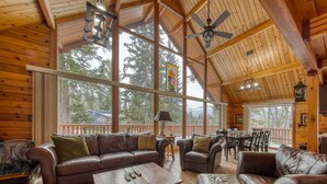 A spacious wooden cabin with large windows, a ceiling fan, and a cozy seating area with leather sofas and a wooden coffee table. - Spring Creek Cabin | 6BR | Hot Tub & Sauna (Fairmont Hot Springs)