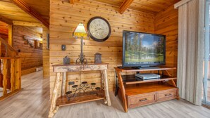 A wooden cabin interior with a wooden TV stand, a clock, and a rustic table. - Spring Creek Cabin | 6BR | Hot Tub & Sauna (Fairmont Hot Springs)