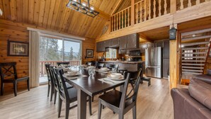 A wooden cabin interior with a dining area, kitchen, and a staircase leading to a loft. - Spring Creek Cabin | 6BR | Hot Tub & Sauna (Fairmont Hot Springs)