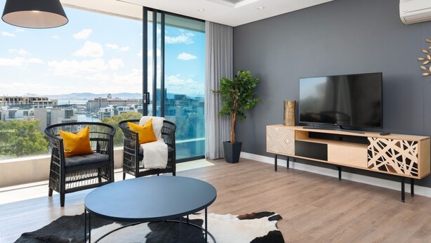 A modern living room with a large TV, comfortable seating, a potted plant, and a city view. - Designer apartment close to the Waterfront (Cape Town)