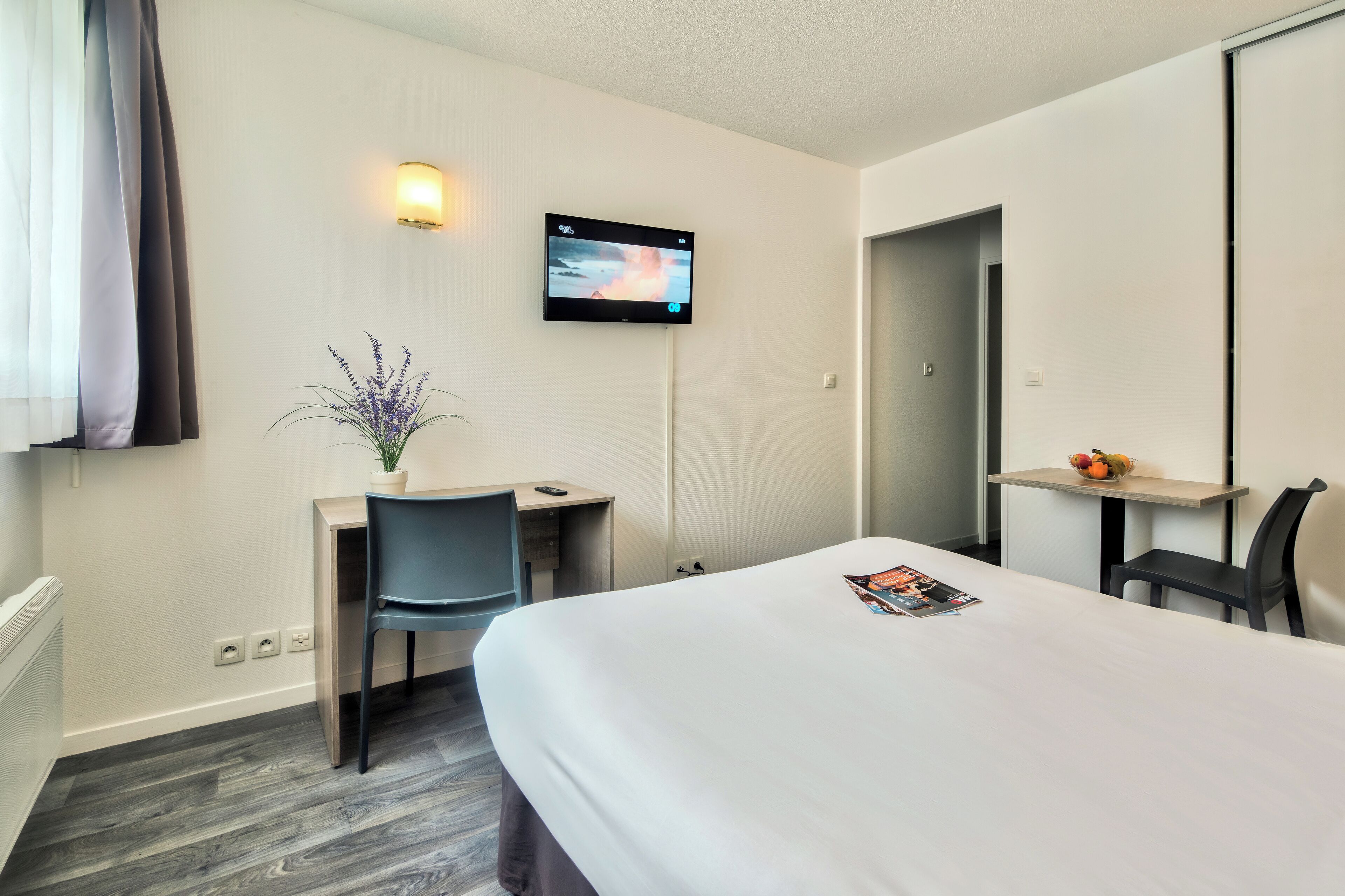 A hotel room with a bed, a desk, a chair, a TV mounted on the wall, and a small table with fruit.
