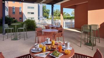 Une terrasse extérieure avec des tables et des chaises rondes, une disposition pour le petit-déjeuner, et une vue sur les bâtiments en arrière-plan.
