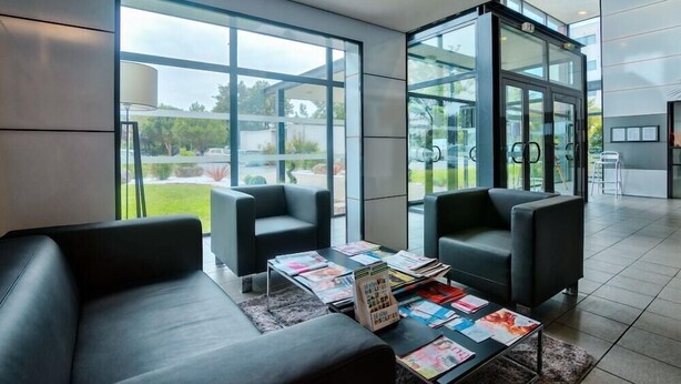 A modern lobby with two black leather sofas, a coffee table with magazines, and large glass doors leading to a garden. - Zenitude Hotel-residences Bordeaux Aeroport Merignac (Merignac)