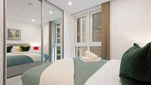 A modern hotel room with a large bed, a mirror, a window with blinds, and a view of the city. - One Bedroom Apartment at Alie Street in Aldgate London by MySquare (London)