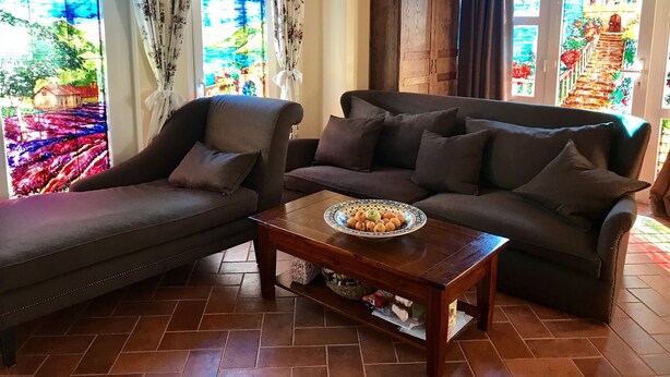 Ein Wohnzimmer mit einer Couch, einem Sessel und einem Couchtisch mit Obst. Es gibt Buntglasfenster und eine Kristallleuchter.
