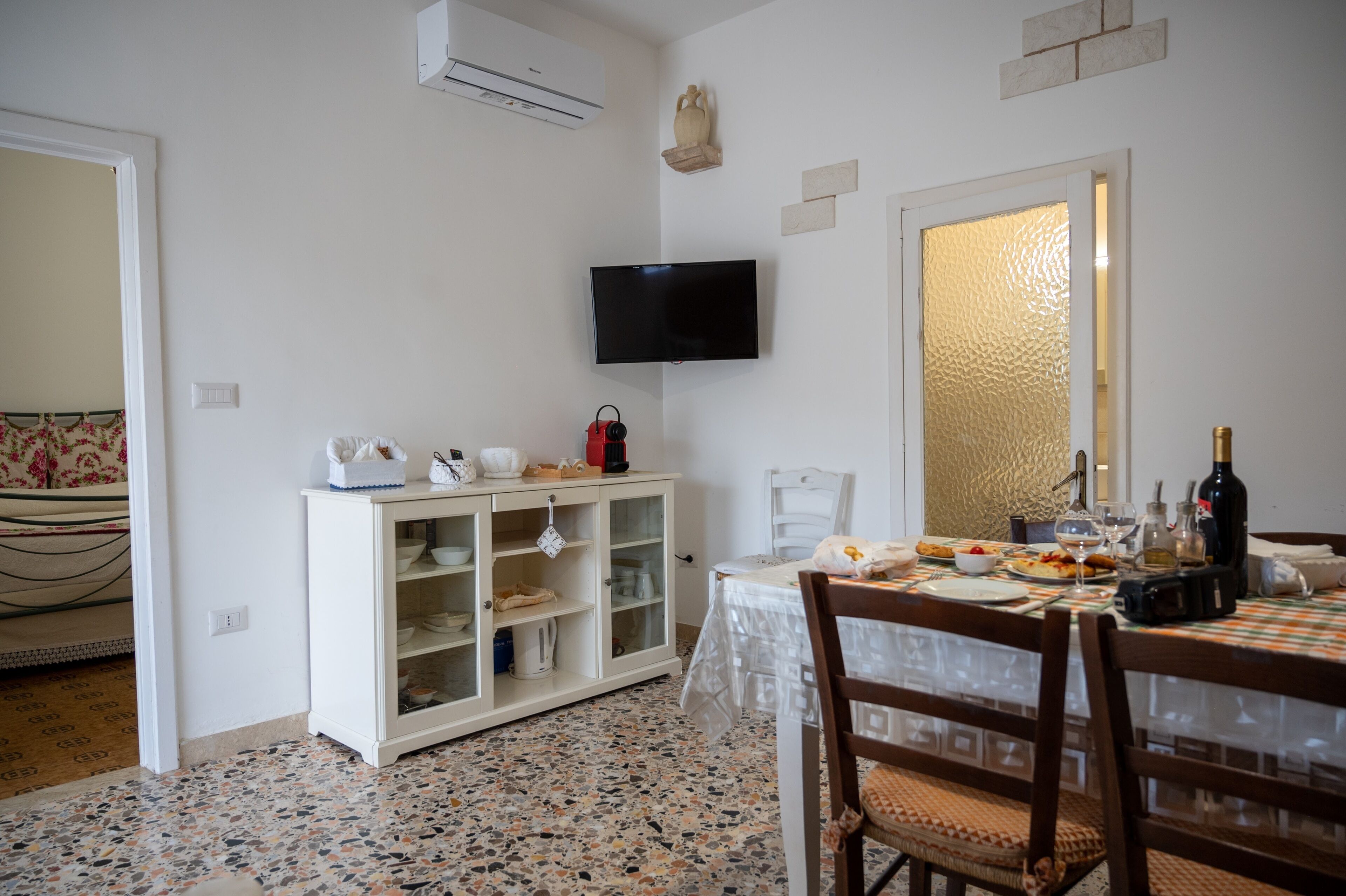 Uma sala de jantar com uma mesa posta para dois, uma televisão fixada na parede e um armário com portas de vidro.