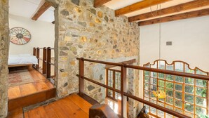 A rustic room with a stone wall, wooden beams, a bed, and a view through a window.