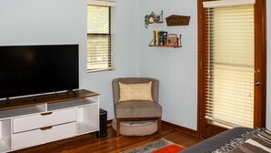 Ein Wohnzimmer mit Flachbildfernseher, grauem Sessel, kleinem Beistelltisch und Bücherregal mit Büchern und einem Vintage-Radio.