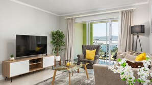 Una sala moderna con un televisor de pantalla plana, una mesa de centro de vidrio, un sofá con cojines amarillos, un sillón grande y vistas de un paisaje urbano a través de la ventana.