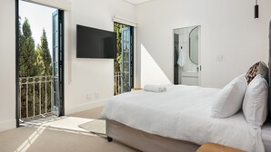 A modern bedroom with a large bed, a TV mounted on the wall, and a balcony with a view of trees. - Modern Apartment in De Waterkant l Private Rooftop (Cape Town)