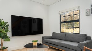 A modern living room with a large flat-screen TV mounted on the wall, a grey sofa, a small round coffee table with a plant, and a window with blinds. - Modern Apartment in De Waterkant l Private Rooftop (Cape Town)