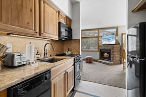 A modern kitchen with wooden cabinets, a stone fireplace, and a large window overlooking a snowy landscape. - Keystone with EPIC Views (Keystone)