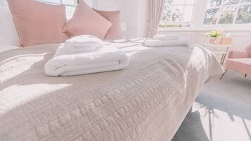 Una cama bien tendida con toallas dobladas, una silla rosa y una ventana con cortinas.