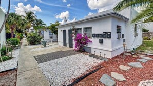 A modern single-story house with a white exterior, a small front porch, and a well-maintained garden with palm trees and purple flowers. - PRIME DELRAY BEACH, CHARMING 2 BEDROOM COTTAGE, PETS WELCOME  (Delray Beach)