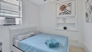 A bedroom with a bed, a ceiling fan, a window with blinds, a wall-mounted shelf with decorative items, and a wall with a floral painting. - PRIME DELRAY BEACH, CHARMING 2 BEDROOM COTTAGE, PETS WELCOME  (Delray Beach)