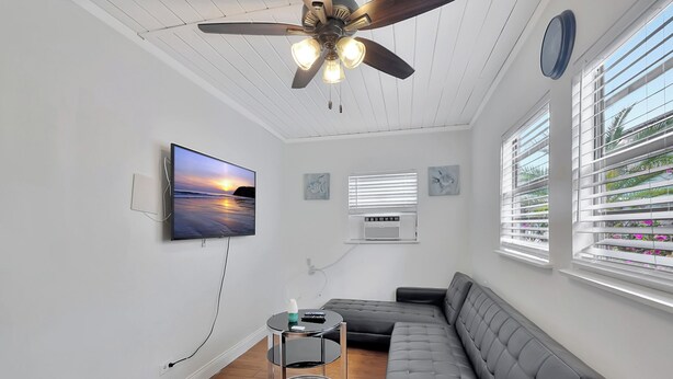 A modern living room with a grey sofa, a ceiling fan, a mounted TV, and a window with blinds. - PRIME DELRAY BEACH, CHARMING 2 BEDROOM COTTAGE, PETS WELCOME  (Delray Beach)