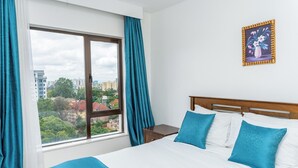 Een hotelkamer met een groot raam, een bed met blauw beddengoed en een ingelijst schilderij aan de muur.