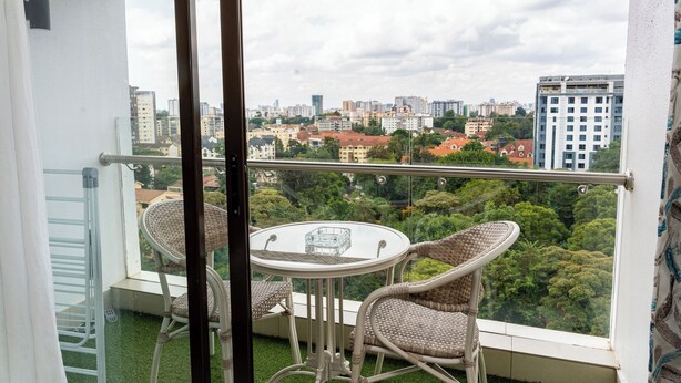 Een balkon met een glazen tafel en rieten stoelen, met uitzicht op een stadslandschap vol gebouwen en groen.