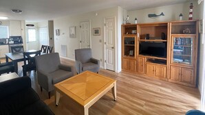 A living room with a wooden coffee table, grey armchairs, a TV stand with shelves, and a kitchen in the background. - Tern Inn - Water Front - Townhouse, deep water dock. LINENS AND TOWELS, (Chincoteague)