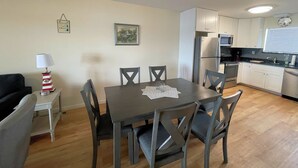 A dining area with a table and chairs, a lamp, a framed picture, and a kitchen with cabinets and appliances. - Tern Inn - Water Front - Townhouse, deep water dock. LINENS AND TOWELS, (Chincoteague)