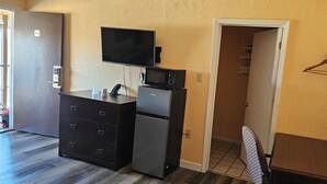 Habitación compacta con kitchenette, un televisor de pantalla plana sobre el microondas, un refrigerador pequeño, un aparador de madera y una puerta que da a un armario con ropa colgada.
