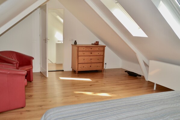 A room with a sloped ceiling, wooden flooring, a red chair, a wooden dresser, and a bed.