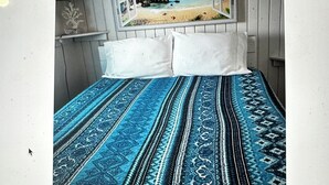 Ein Bett mit blauer, gemusterter Tagesdecke, zwei weiße Kissen und ein Fenster mit einer Strandmotiv-Szenerie.