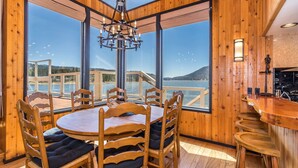 Un interno in legno con un'ampia finestra vista su un lago e delle colline. Si nota un tavolo rotondo in legno con sedie, un angolo bar e un lampadario.
