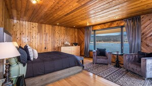 Camera da letto in una baita di legno con un letto grande, una cassettiera, un angolo salotto con un tavolo e vista su un lago.