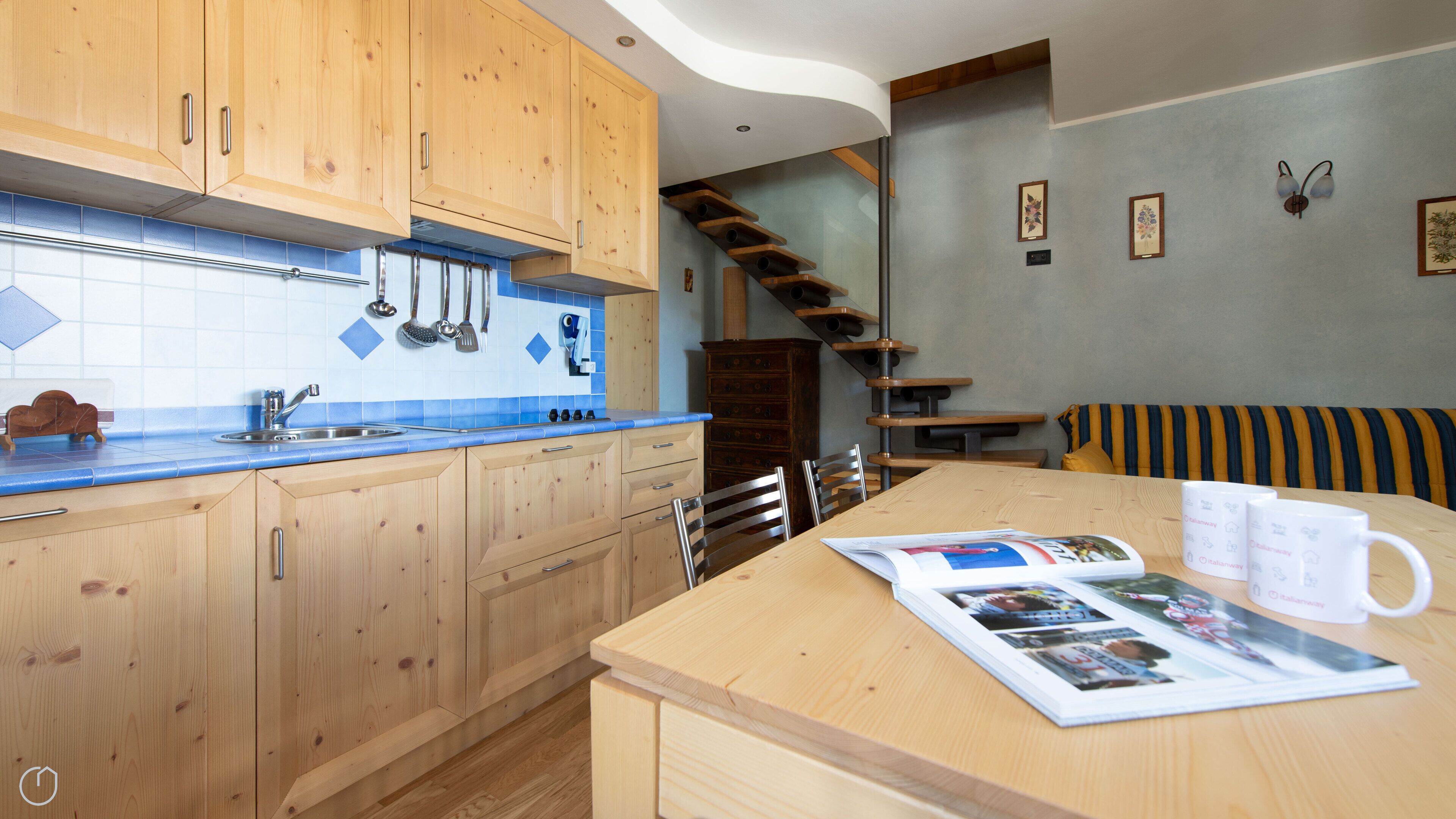 Een moderne keuken met houten kastjes, een blauwe betegelde achterwand en een houten eettafel met tijdschriften en een koffiemok.