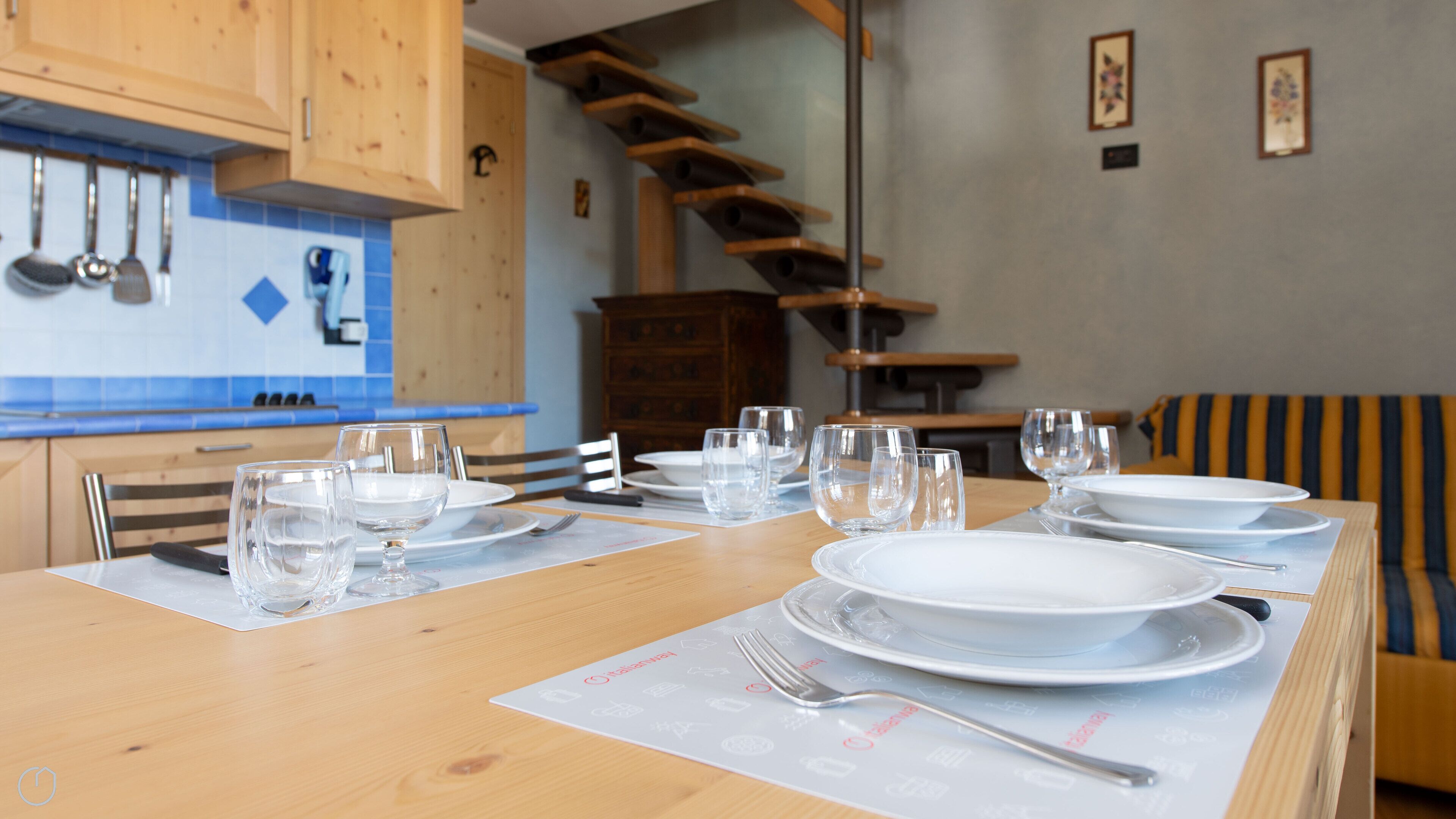 Een eettafel gedekt met borden, glazen en bestek in een keuken met houten kasten en een blauwe tegelachtergrond.