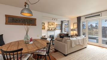A cozy living room with a fireplace, a dining area with a wooden table, and a sofa with a view through large windows.