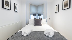 Una cama bien hecha con dos almohadas, una colcha y dos toallas enrolladas. Hay cuadros en las paredes y una ventana con cortinas.