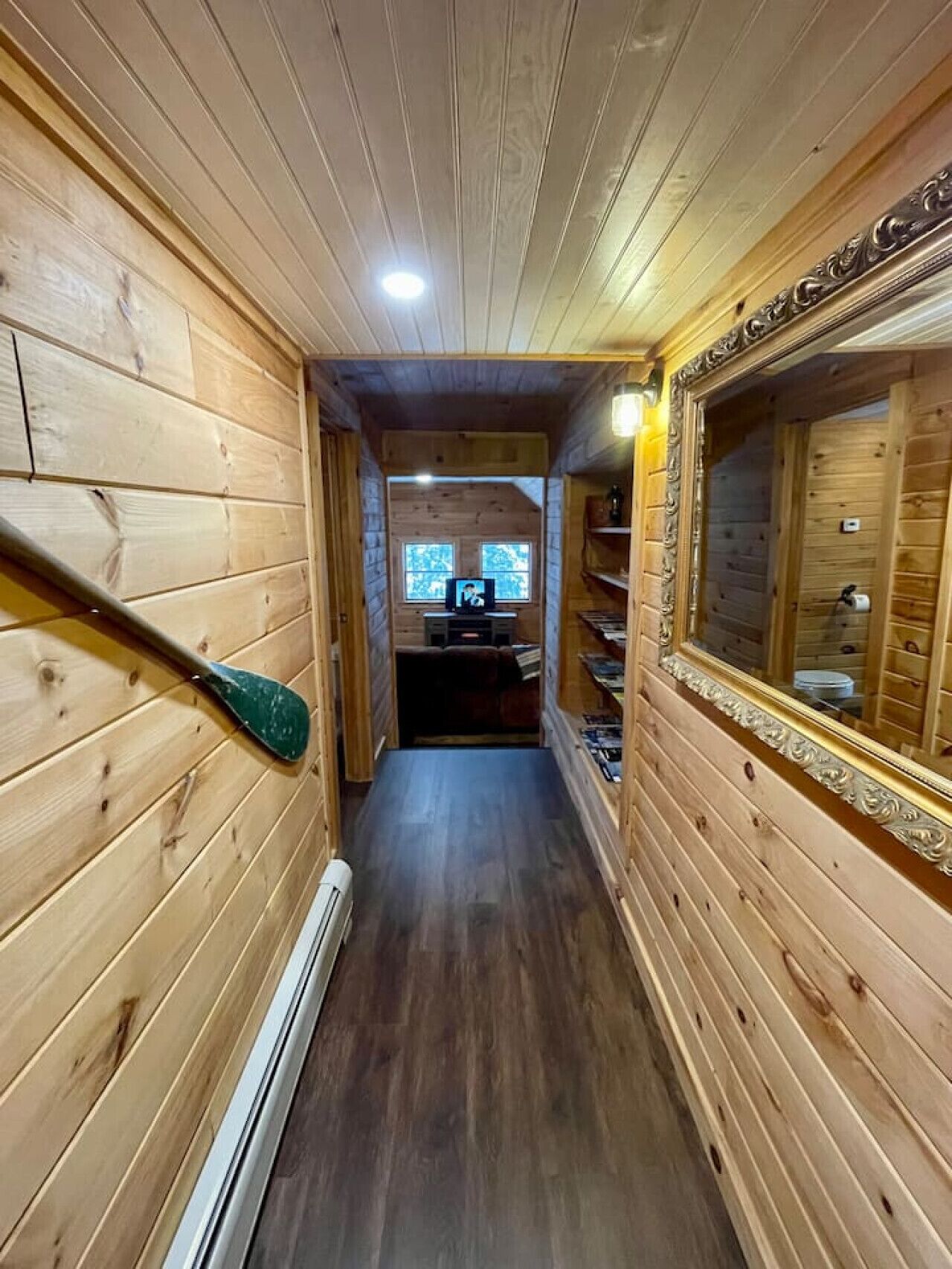 A narrow wooden hallway with a paddle leaning against the wall, a mirror on the right, and a television in the background.