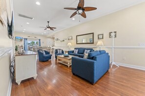 A spacious living room with a large blue sofa set, a coffee table, and two ceiling fans. - Fun In The Sun, Luxury Beach House with Pool, Blocks from Beach (North Myrtle Beach)