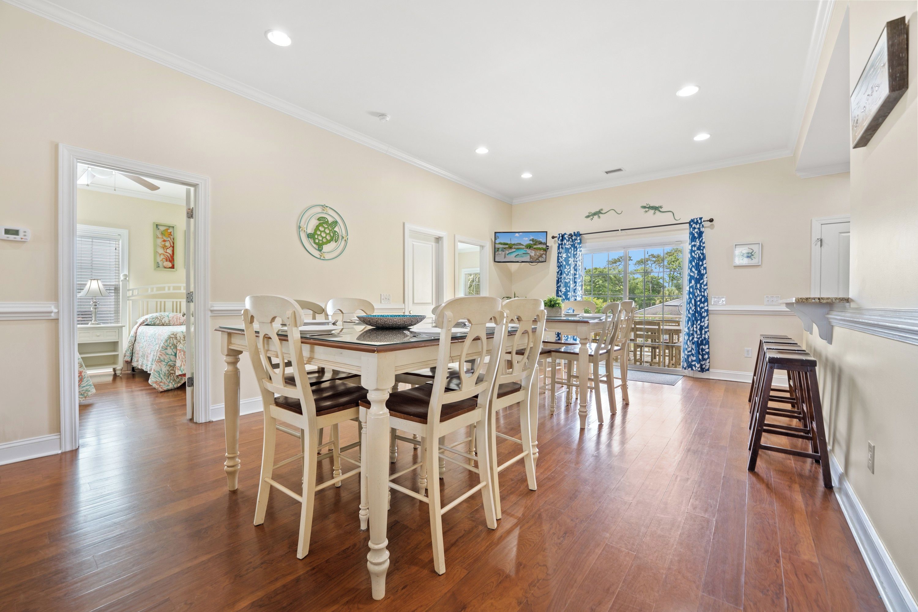 A dining area with a wooden table and chairs, a television mounted on the wall, and a view of the outdoors through a window.