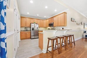 A spacious kitchen with wooden cabinets, a stainless steel refrigerator, a microwave, and a granite countertop. There are three wooden stools at the island. - Fun In The Sun, Luxury Beach House with Pool, Blocks from Beach (North Myrtle Beach)