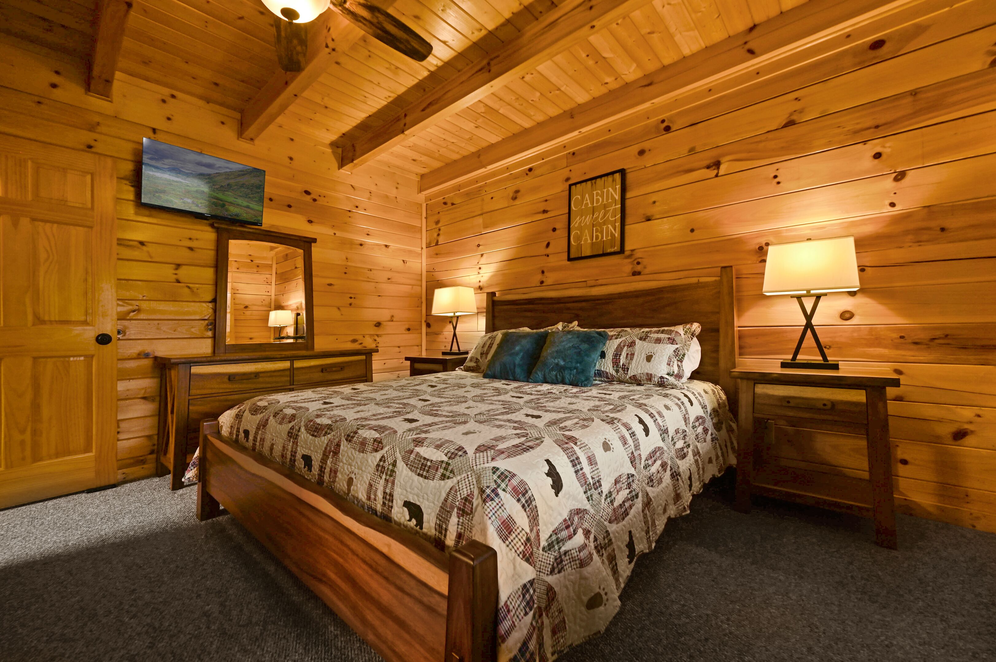 A wooden cabin bedroom with a bed, two nightstands, a mounted TV, and a framed sign on the wall.