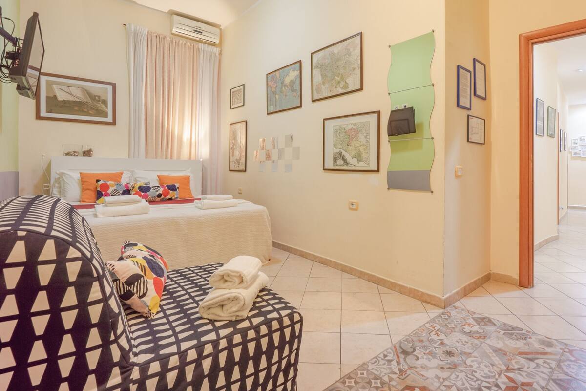 Quarto de hotel com cama, sofá, mesa pequena e parede com quadros.