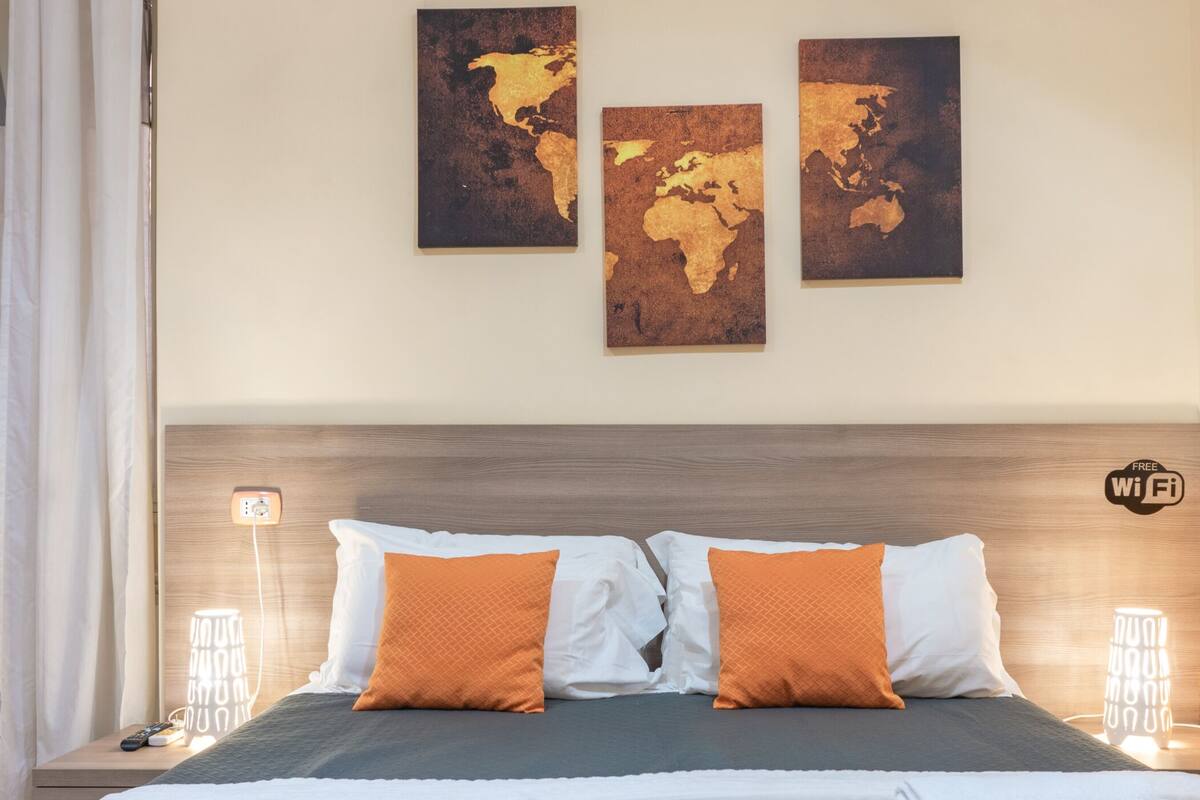 Quarto de hotel com cabeceira de madeira, cama com almofadas brancas e laranja, e três obras de arte com mapas-múndi na parede.