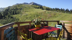 Um deck de madeira com uma mesa vermelha e cadeiras amarelas, com vista para uma paisagem montanhosa.