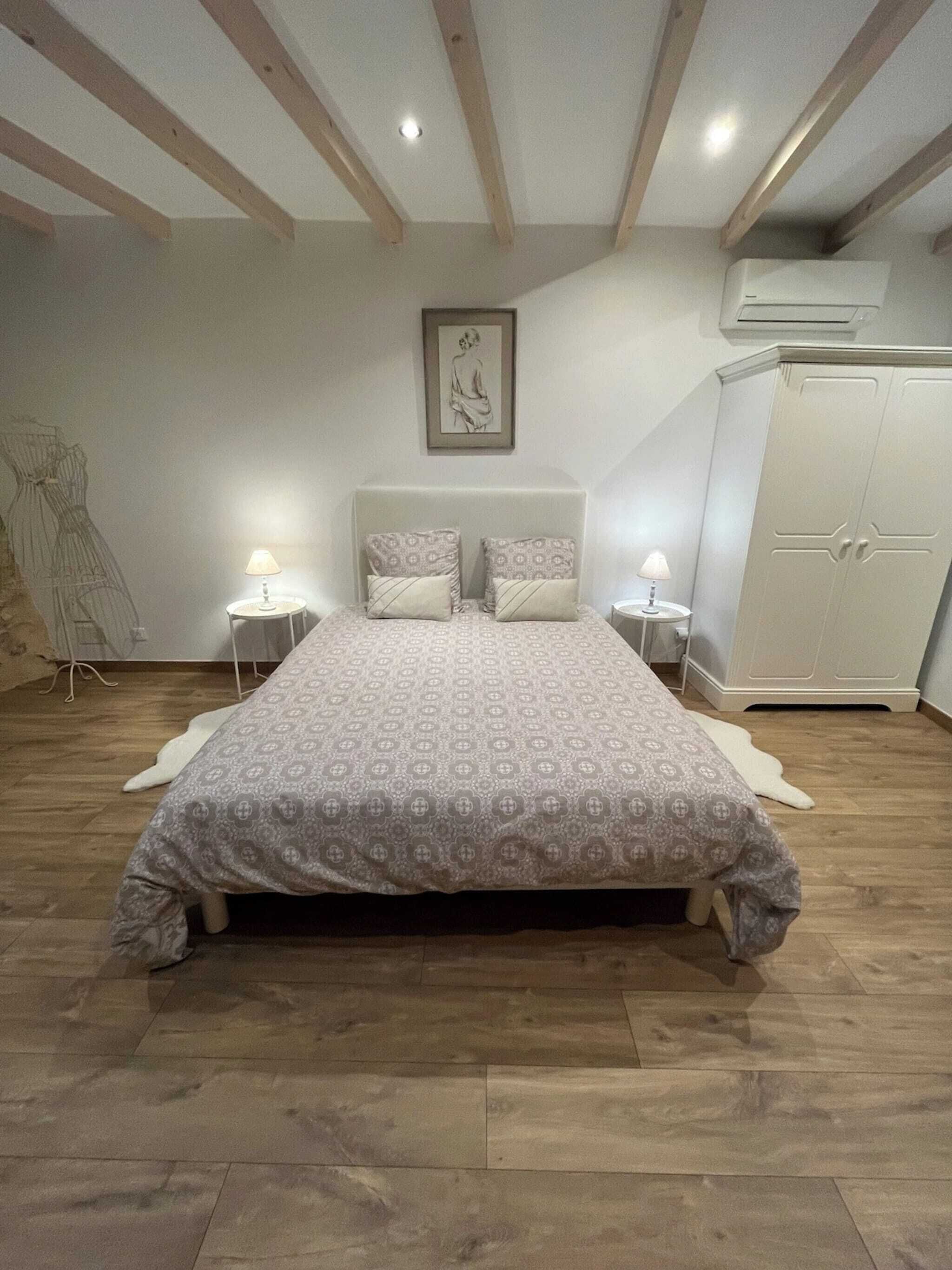 A bedroom with a large bed, two bedside tables with lamps, a wardrobe, and an air conditioning unit on the wall.