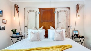 Quarto com cama grande, duas mesas de cabeceira, porta de madeira e luminárias fixadas na parede.