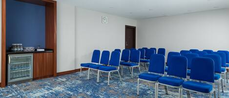 A conference room with rows of blue chairs, a refrigerator with water bottles, and a clock on the wall.