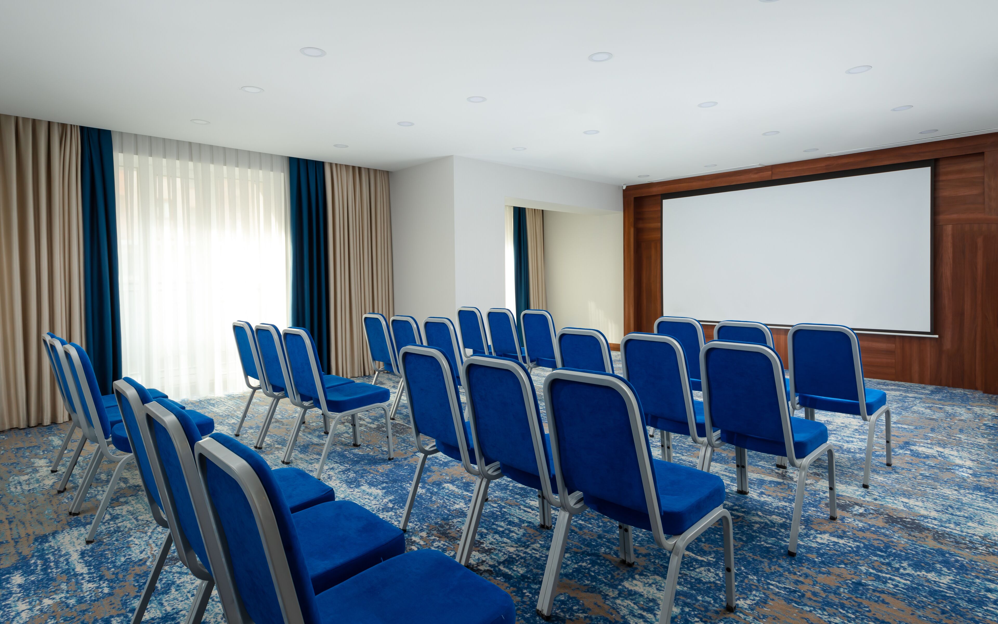 A conference room with rows of blue chairs, a white projection screen, and a carpeted floor.