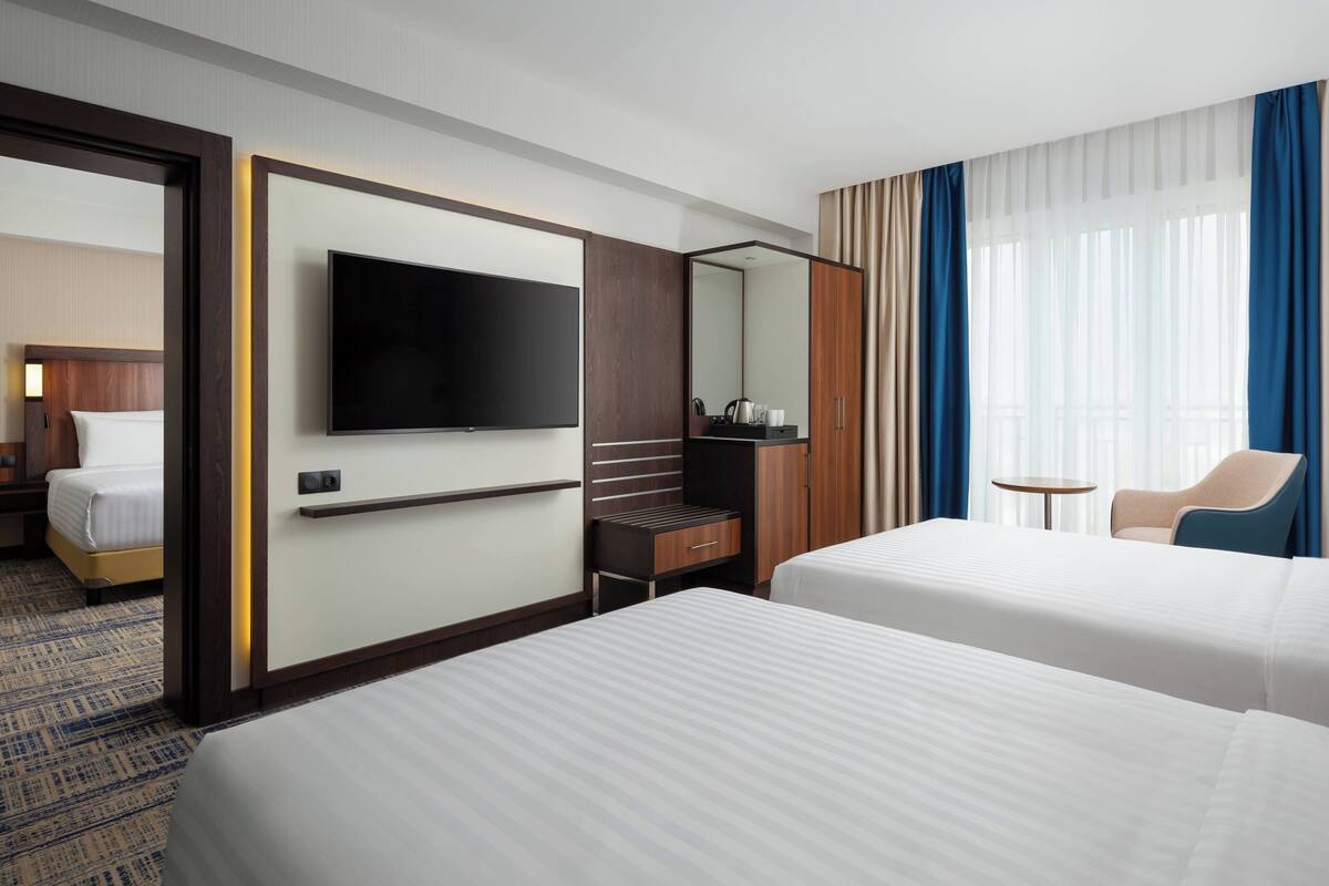 A hotel room with two beds, a television, a desk with a chair, a small table, and a window with curtains.